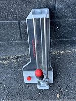 Top view of the gray metal tile cutter showing the parallel metal rails and red adjustment knob at the bottom left.