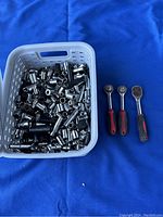 Lot of over 100 metal sockets placed in a white plastic basket with three ratchet handles displayed for scale.