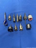 Top-down view of 11 concrete hole drill bits of mixed sizes on blue surface showing various cylindrical hole saw bits with teeth and a blue-coated masonry bit