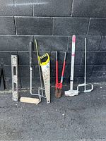 Photo of 7 hand tools arranged against black wall showing various tools like a level, paint roller, saw, bolt cutters, paint mixer, sledgehammer, and welding clamp.