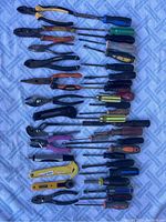 Photo showing an assortment of pliers, wire cutters, and screwdrivers arranged on a patterned cloth background. Handles vary in color and material.