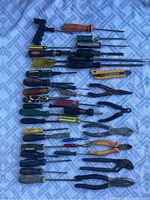 Top down view of over 40 assorted construction hand tools spread on a blue textured fabric. Visible tools include multiple screwdrivers with different handle types, several pliers, wire cutters, and a coping saw with a wooden handle.
