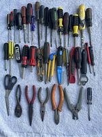 Top down view of over 40 hand tools arranged on a light-colored fabric surface including multiple screwdrivers, pliers, and scissors.