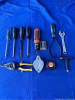 Top-down view of all 12 hand tools arranged on blue fabric showing variety including screwdrivers, wrench, tape measure, tin snips, chalk line reel, and strap wrench.