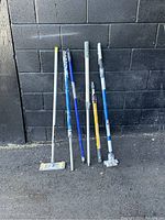 Lot of six construction taping tools with handles against black brick wall, showing variety of tool heads and handle colors.