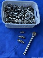 Photo showing white basket filled with over 100 chrome sockets, two ratchet handles and three universal socket adapters placed in front on a blue surface.