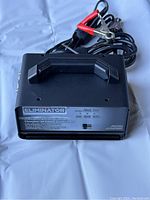 Black Eliminator battery charger with handle and attached red and black alligator clamp cables shown from top side.