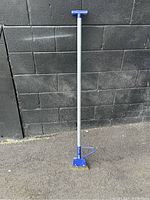 Floor scraper standing upright against a black brick wall, showing full length and handle detail.