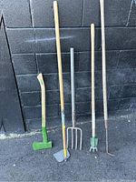 Five garden tools including a sledgehammer, hoe, garden fork, cultivator, and weeding tool against a black brick wall.