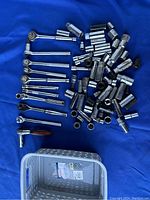 Over 70 pieces of chrome socket wrenches, ratchets, and extensions arranged on blue fabric with a perforated gray plastic storage box visible.