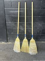 Three brand new outdoor brooms with natural bristle heads and wooden handles, standing against a black brick wall. Each broom’s bristle head is wrapped in protective plastic.