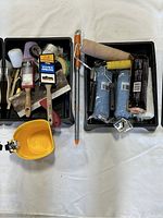 Top-down view of two black trays containing assorted painting tools including rollers, brushes, scrapers, and a yellow paint container.