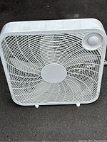 Front view of the white plastic box fan on pavement showing the grille, blades, and speed control knob.