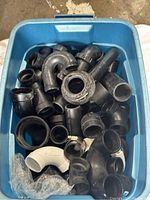 Top view of blue plastic bin filled with black plastic plumbing fittings including elbows, connectors, couplers, and adapters.
