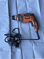 Side view of the Ridgid R7001 corded drill lying flat on a gray surface with power cord coiled beside it.