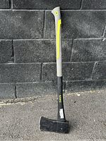 Full view of the Yard Works axe standing against a black brick wall on asphalt ground, showing the entire tool including handle and axe head.