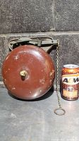 Front view of the antique Bevin Bell showing the round bell and pull chain next to an A&W soda can for scale.