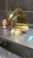 Side view of brass plated sailboat mounted on crystal stone base placed on a metal surface, with an A&W soda can and lighter in the background for size reference.