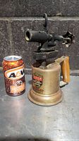 Side view of the antique brass blow torch placed next to an A&W soda can for scale, showing the brass tank base, wooden handle, and metal nozzle with adjustment valve.
