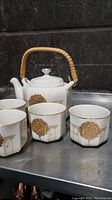 Photo showing white ceramic teapot with bamboo handle and four hexagonal tea cups with gold floral tree designs.