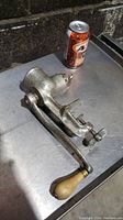 Photo showing the manual meat grinder with a wooden handle placed on a metal surface next to a soda can for scale.
