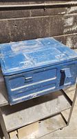 Front and right side angle view of blue painted metal military storage crate on a metal shelf against a concrete block wall showing hinged lid, latch closures, and carry handle.