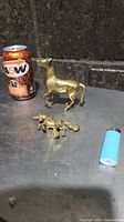 Two miniature brass figurines photographed on a metallic surface next to an A&W soda can and a lighter for size reference.