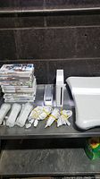 Photo showing the white Nintendo Wii console, three Wii remotes with wrist straps, three Nunchuk controllers, and a stack of game cases