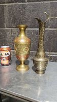 Two brass vases displayed on metal surface against a concrete wall, alongside an A&W soda can for size comparison. One vase is shorter with red ornamental designs, the other is taller with a handle and floral patterns.