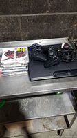 Front view of PS3 console with 2 black controllers, 7 PS3 games stacked beside them and AV cable.