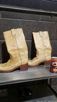 Side view of both boots showing beige leather and wooden heels with some discoloration, placed on a metal shelf with a soda can for size reference.