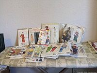 Photo showing 12 vintage sewing patterns arranged on a table, featuring various clothing styles including dresses, coats, and jumpsuits in illustrated vintage packaging.