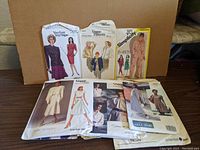 Photo showing 7 sewing pattern envelopes arranged upright and fanned out; includes patterns from Vogue, Simplicity, Very Easy, John Anthony brands with visible model illustrations for dresses, jackets, and suits.