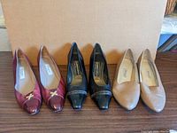 Three pairs of ladies shoes lined up side by side showing colors and styles: burgundy with cross hardware, black with chain detail, beige plain style.