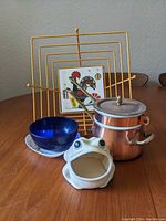Lot displayed on wooden table showing all items together: gold-tone metal toast rack, white ceramic frog container, blue glass bowl, copper pot with lid, and rooster tile coaster.
