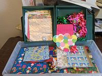 Green plastic container with assorted gift bags and multiple sheets of Christmas wrapping paper flat inside, featuring various holiday patterns and colors.