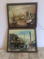 Photo of two framed paintings stacked vertically on a fabric surface, showing both harbor scenes with sailboats and rustic buildings