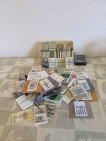 Multiple packages of sewing machine needles and some presser feet with various brand labels and sizes on a table.