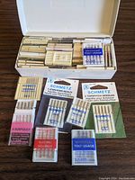 Top view of white plastic storage box opened showing an organized collection of multiple small cases of sewing needles, including Schmetz brand various types.
