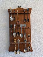 Full view of the wooden display rack with 15 various souvenir spoons hung in rows. Spoons feature different metal finishes, decorative handles, enamel crests, and painted details. Condition shows use and vintage wear.