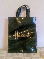 Front view of the dark green shiny plastic bag with gold Harrods logo and handle
