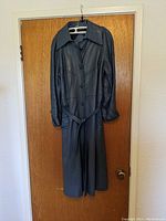 Full view of the black leather ladies coat hanging on a door, showing its length, buttons, belt, and collar.