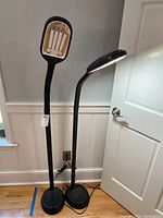 Photo showing two matching black grow lights with flexible necks standing near a wall and door on wooden floor.
