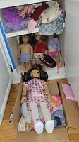 Overview of American Girl dolls and multiple clothing pieces, showing one doll lying in a box and others standing with some garments.