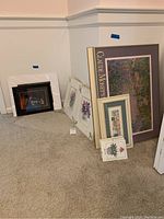 Photo showing ten framed pieces of wall art leaning on a wall including a large Claude Monet poster, floral art prints, and cityscape prints, all on carpeted floor.