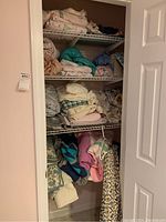 Wire shelving inside closet with stacks of assorted bed linens including sheets, pillowcases, blankets, and quilts in multiple colors and patterns.
