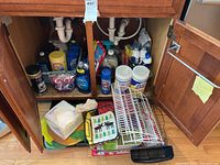 Photo showing cleaning supplies and kitchen accessories stored under a wooden cabinet including Lysol wipes, Dawn dish soap, Cascade dishwasher pods, Easy Off spray, and dish rack with trays and baskets.