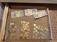 Overview photo showing three groups of coins and three folded banknotes arranged in a cardboard box: English coins on the left, Euros in the middle, wheat penny with some other coins on the right, and three banknotes placed behind them.