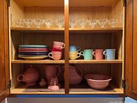Full cabinet shelf showing Fiesta Ware dinner plates, mugs, and glass ice cream dishes on upper shelf