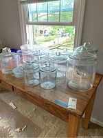 Photo of multiple clear glass canisters with lids placed on a wooden table near a window. Canisters vary in size from small to large.